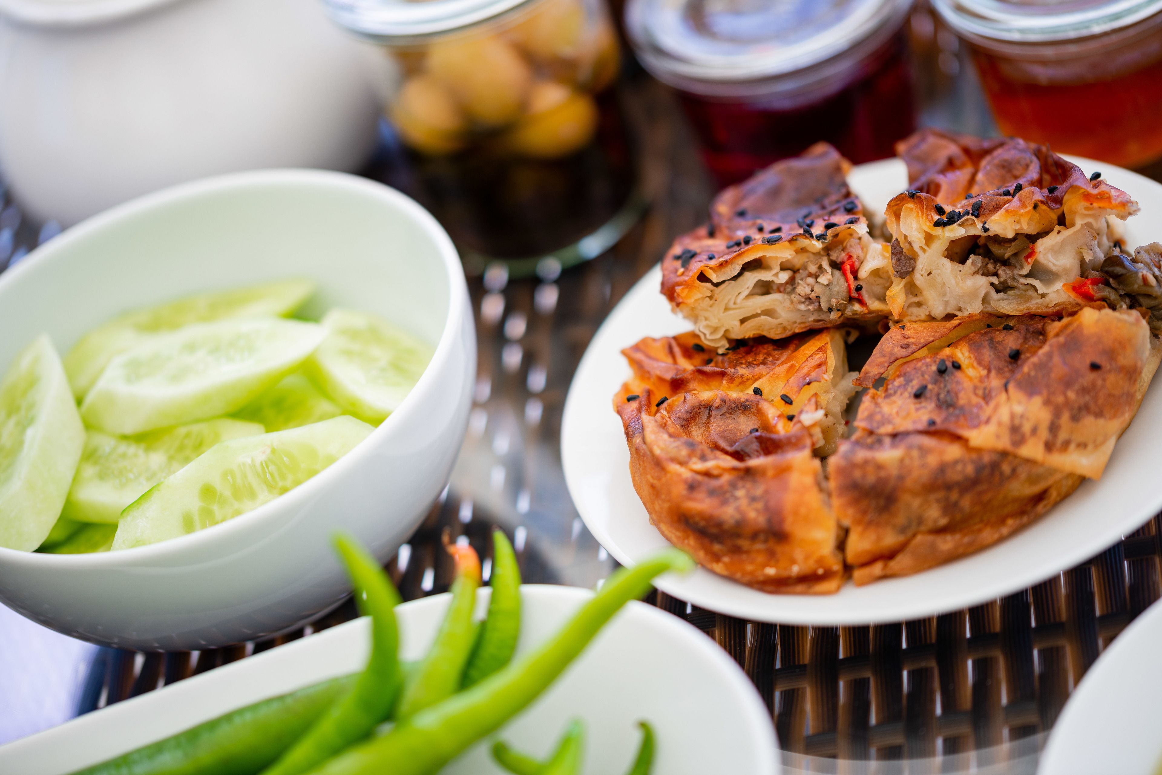 A table with a plate of stuffed pastries, a bowl of sliced cucumbers, and green peppers.
