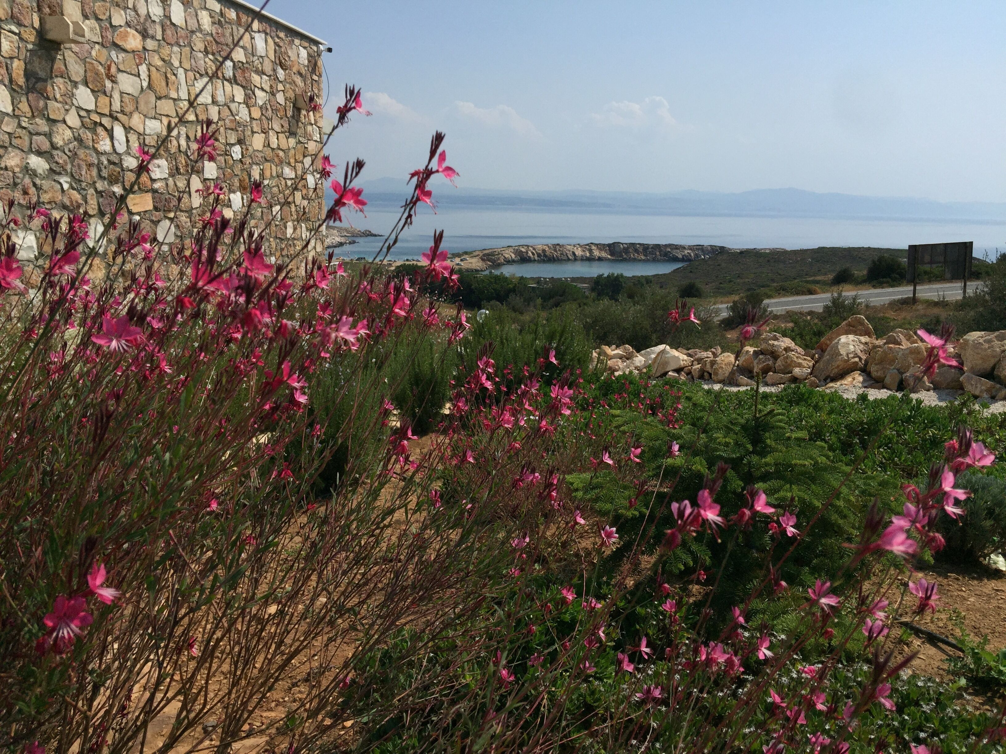 Un edificio de piedra con flores rosadas en primer plano y vistas al mar y a las montañas al fondo.