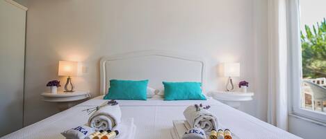 A hotel room with a neatly made bed, two side tables with lamps, and a view of greenery outside.