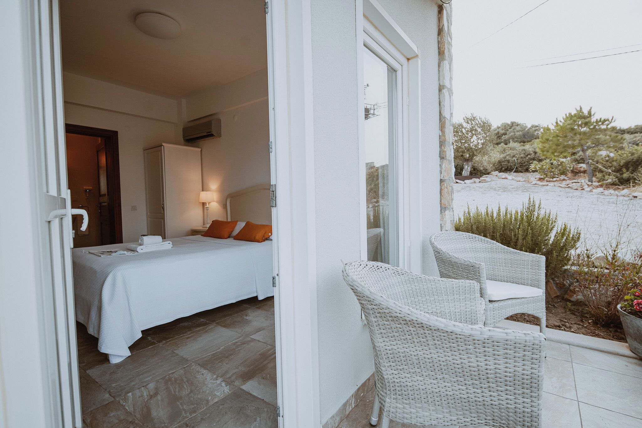 A bedroom with a bed, a nightstand, and a lamp. There is a balcony with wicker chairs and a view of the landscape.