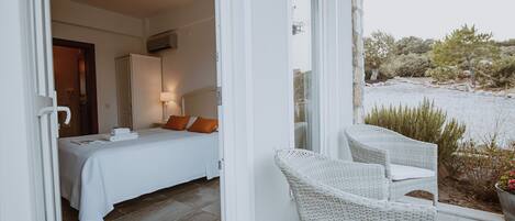 A bedroom with a bed, a nightstand, and a lamp. There is a balcony with wicker chairs and a view of the landscape.