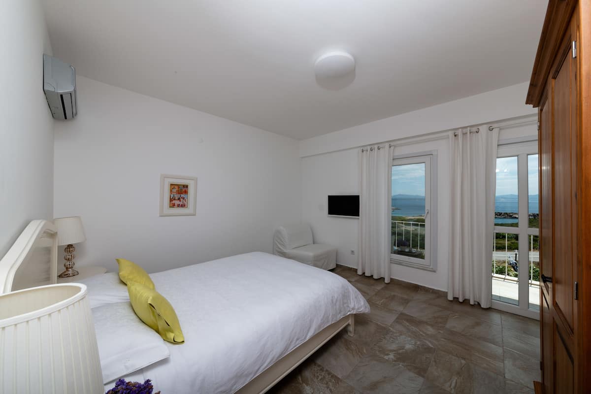 A bedroom with a bed, a chair, a TV, and a balcony with a view.