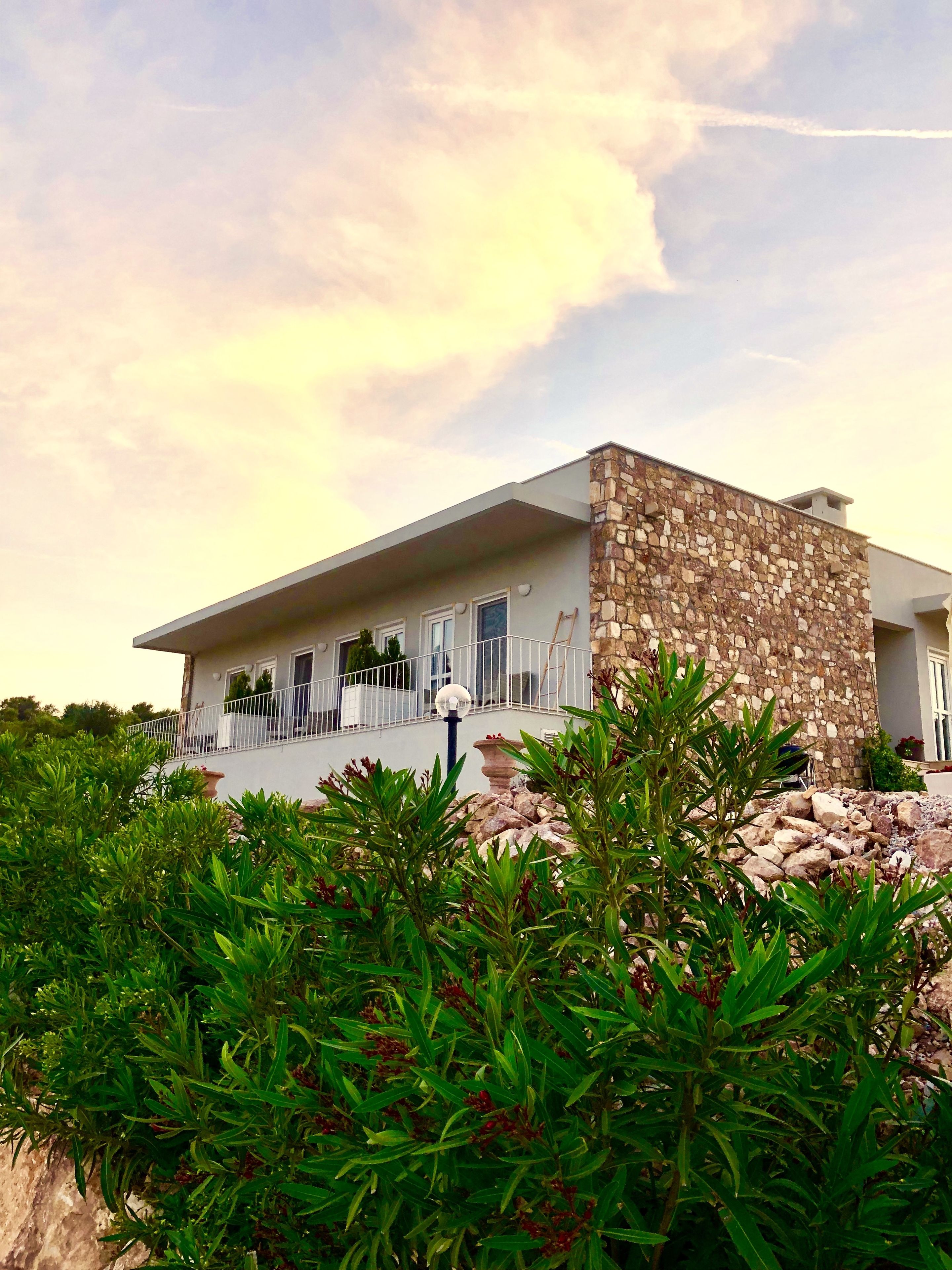 A modern house with a stone facade, a balcony, and lush greenery in the foreground.