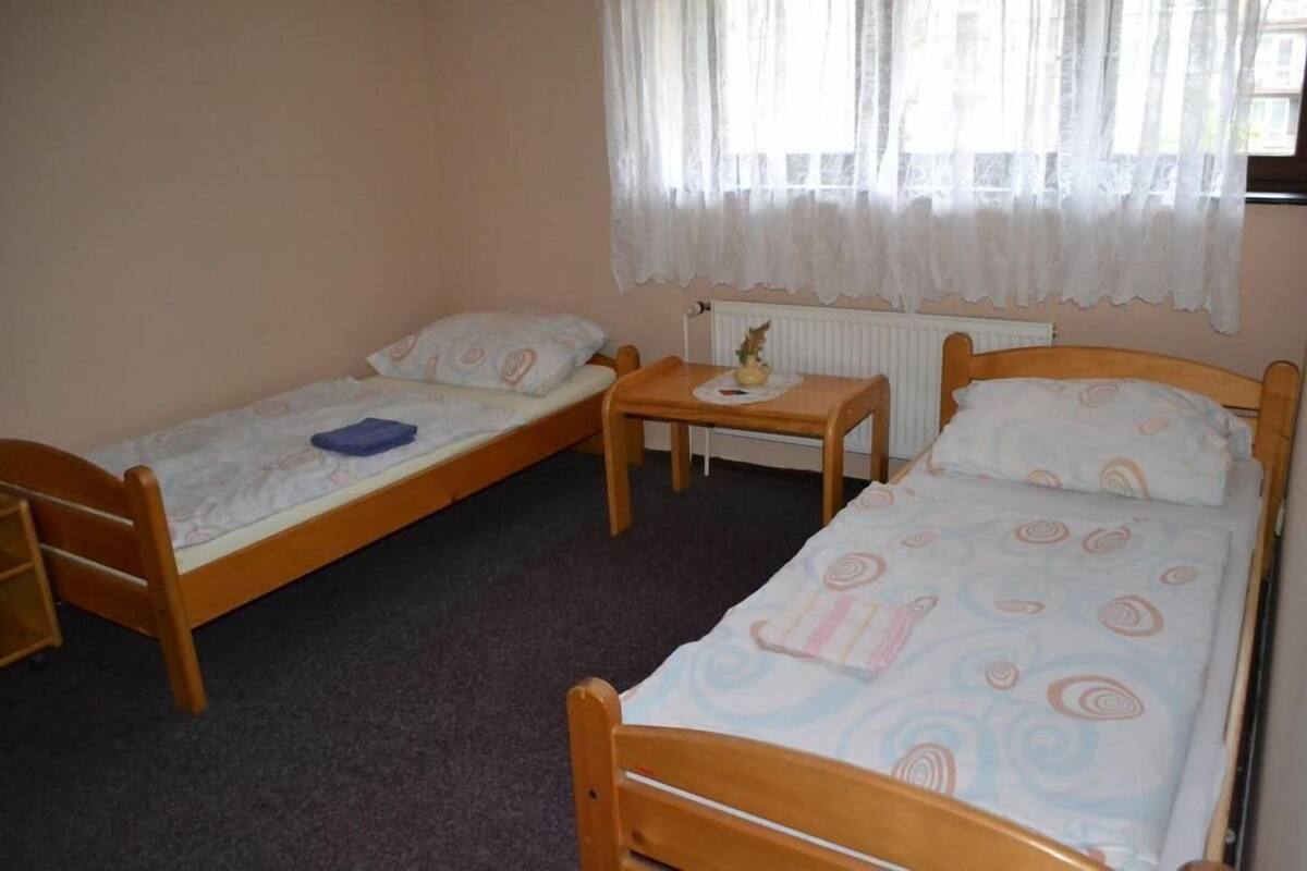 A room with two single beds, a small table, a radiator, and a window with sheer curtains.