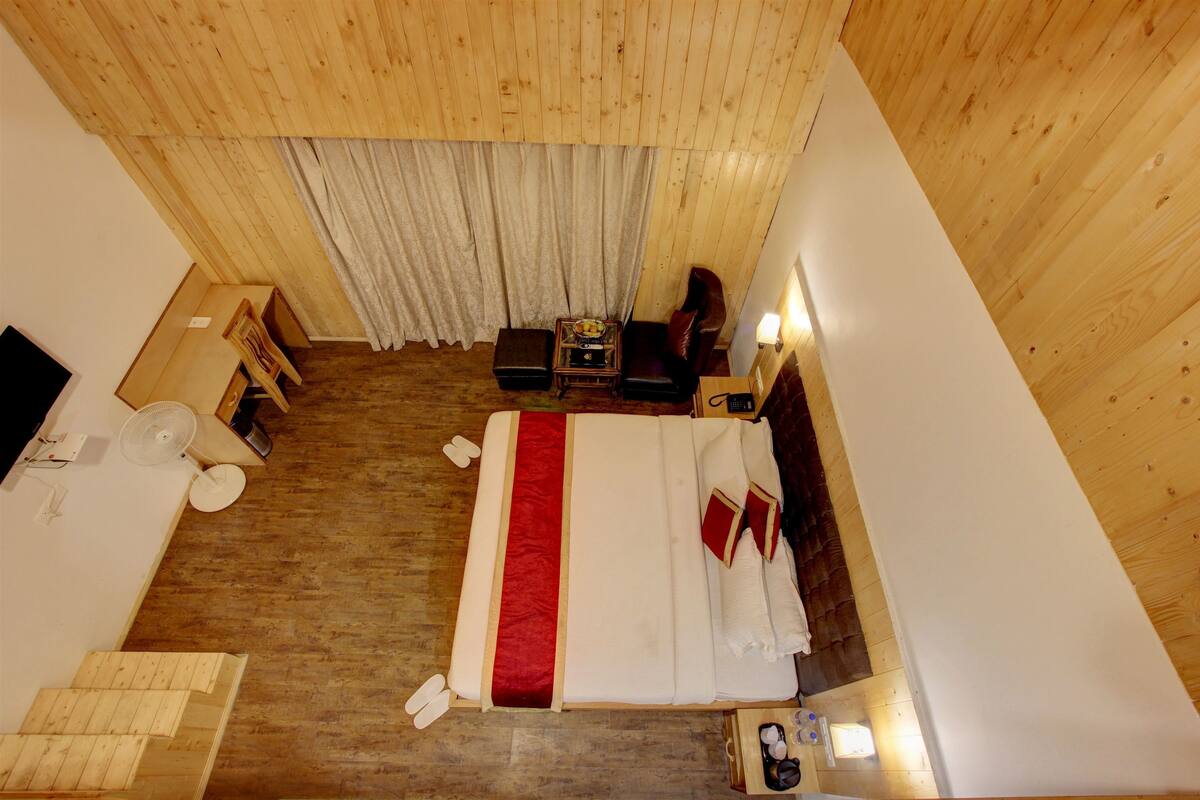 A hotel room with a wooden ceiling, a bed with white bedding and a red accent, a small table with a lamp, a chair, and a window with curtains.
