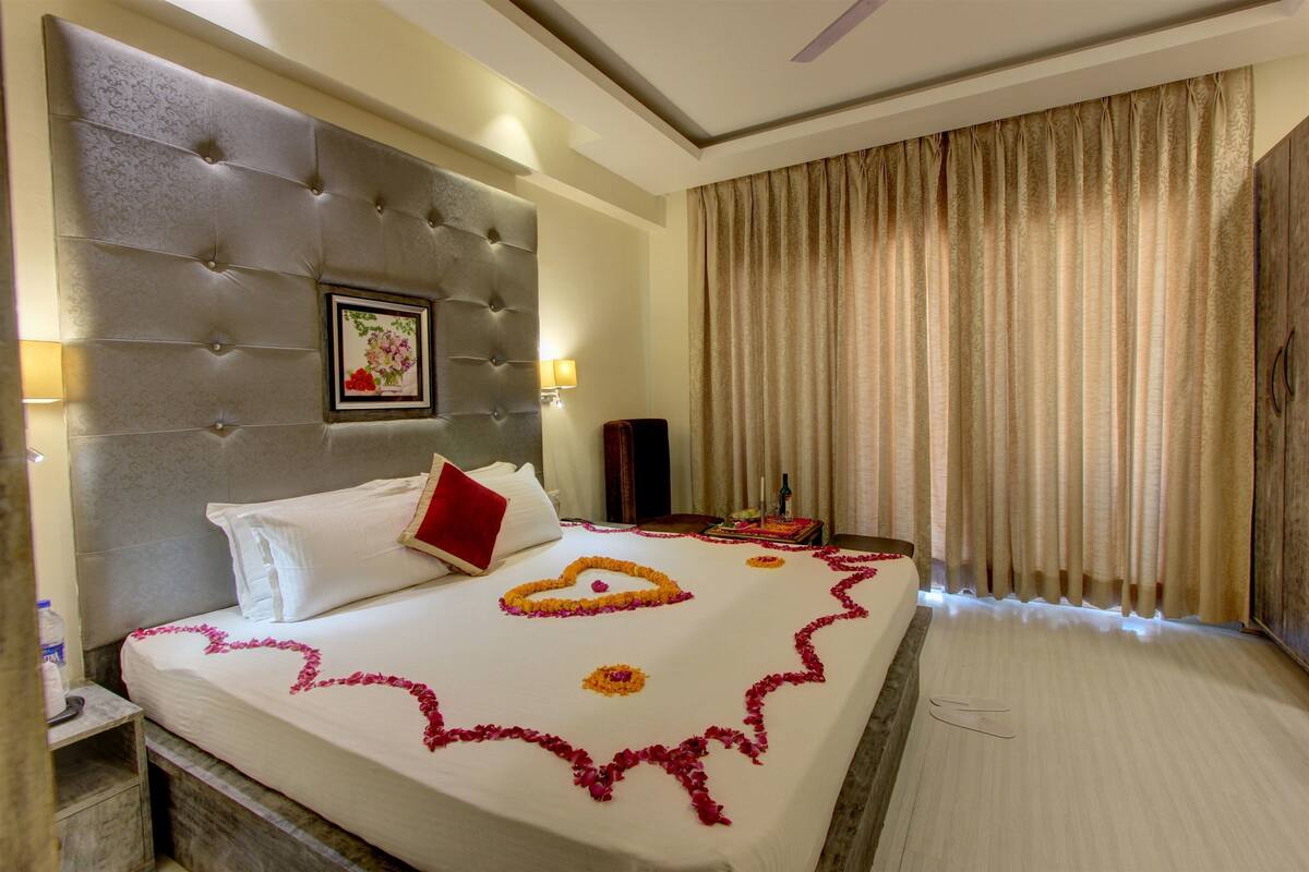 A hotel room with a tufted headboard, a bed with a white bedspread and rose petals, a red pillow, a framed picture on the wall, a speaker, and a large window with curtains.