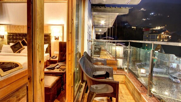 A balcony with a wooden floor, a wicker chair, and a glass railing. - Hill County Resort & Spa, Manali (Manali)