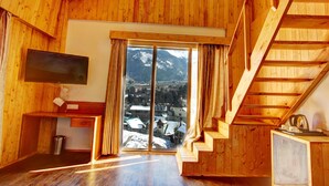 A wooden cabin interior with a TV, a desk, a window with a view of snow-covered mountains, and a wooden staircase. - Hill County Resort & Spa, Manali (Manali)