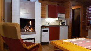 A cozy room with a fireplace, a dining table, and a kitchen area with red cabinets and a microwave. - Seita Cabins (Kolari)