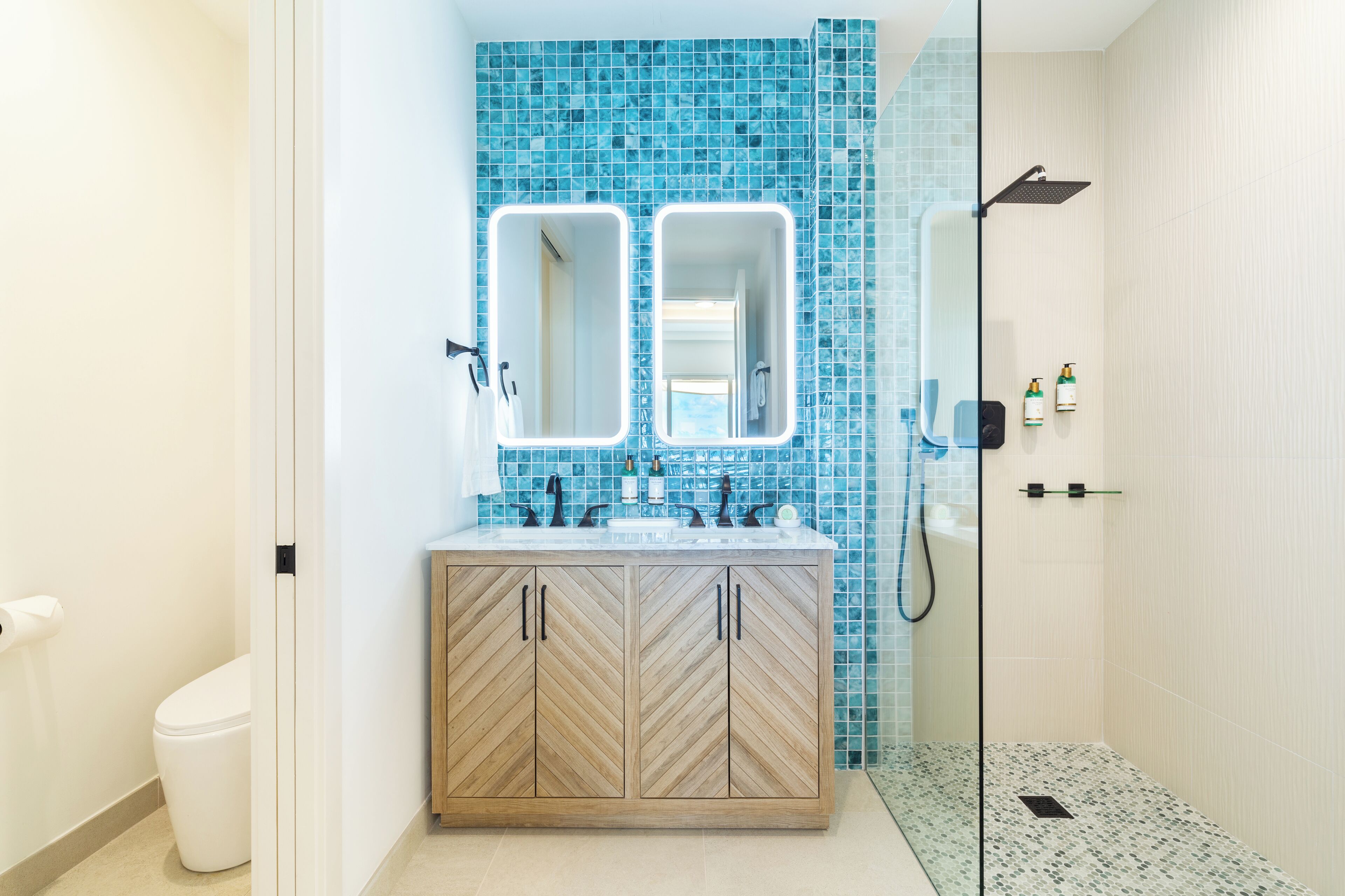 Une salle de bain avec un meuble-lavabo en bois, deux miroirs, une douche et un mur recouvert de carreaux.
