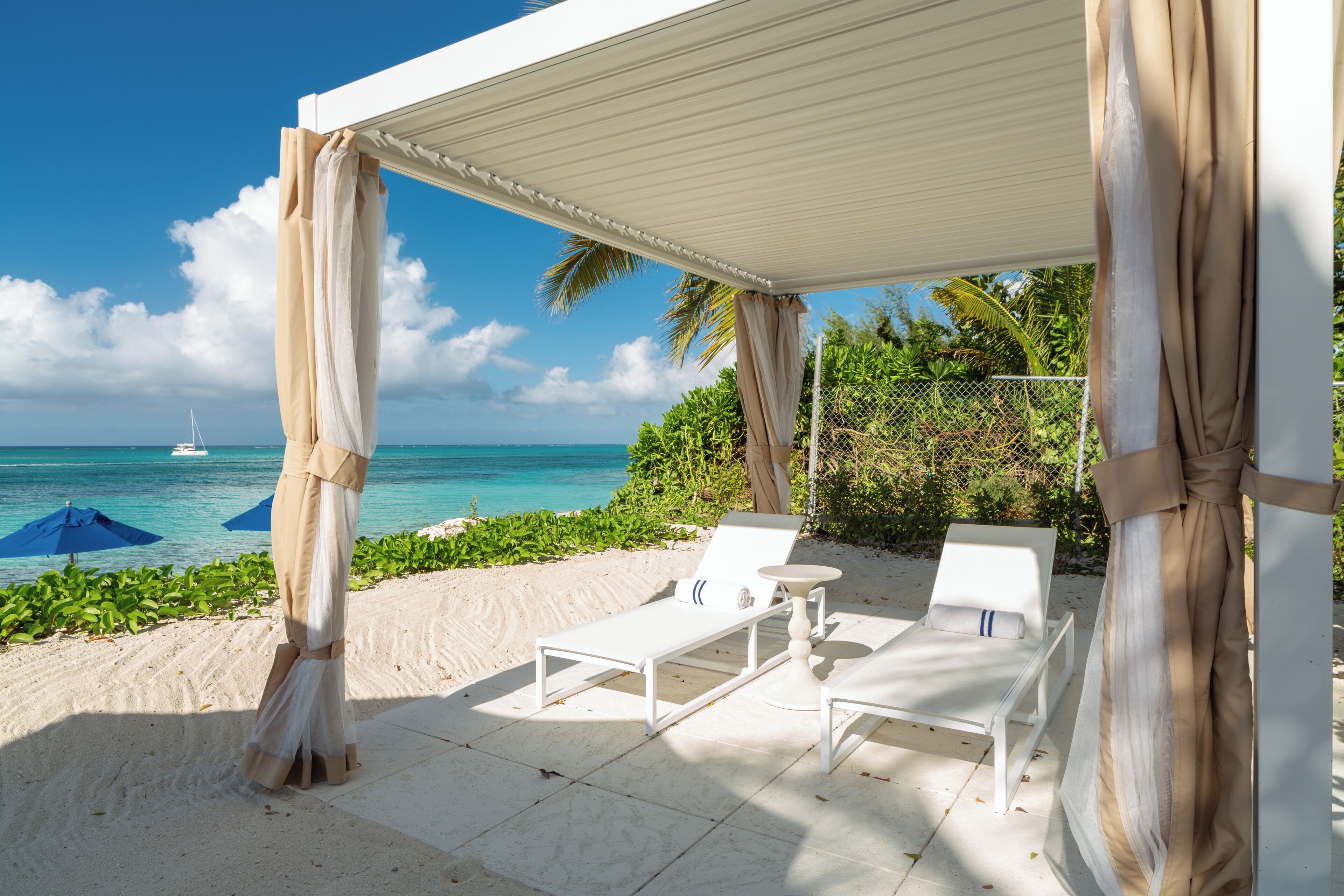 A beachside patio with white lounge chairs and a table, overlooking the ocean with a sailboat in the distance.