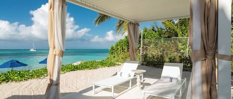 A beachside patio with white lounge chairs and a table, overlooking the ocean with a sailboat in the distance.