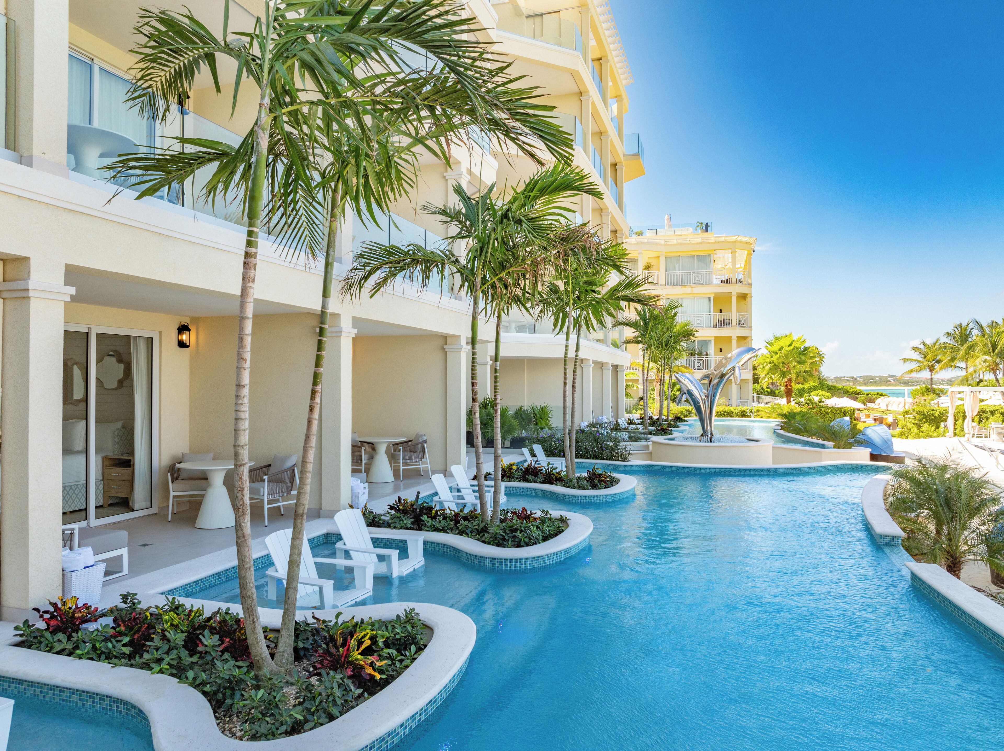 A resort pool area with lounge chairs and palm trees, surrounded by a multi-story building with balconies.