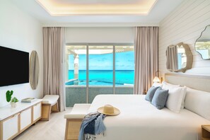 A bedroom with a large bed, a TV, a bedside table, a bench, and a view of the ocean. - Beachfront at Windsong on the Reef (Providenciales)
