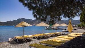 A beach with rows of sun loungers and umbrellas, a boat docked near the shore, and mountains in the background. - Umut Otel & Beach (Marmaris)