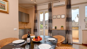 Un comedor con una mesa dispuesta para cuatro, un cuenco de fruta y una botella de vino. Se observan sillas de mimbre y una cocina al fondo.