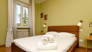 Una cama bien hecha con sábanas blancas y toallas enrolladas, cabecera de madera, mesita de noche con lámpara y ventana con cortinas.