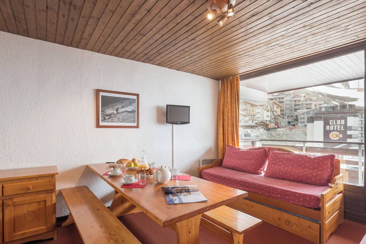 Ein gemütliches Zimmer mit Holzbalkendecke, einer Couch mit roten Kissen, einem Holztisch mit Frühstücksutensilien, einem gerahmten Bild und einem Fernseher.