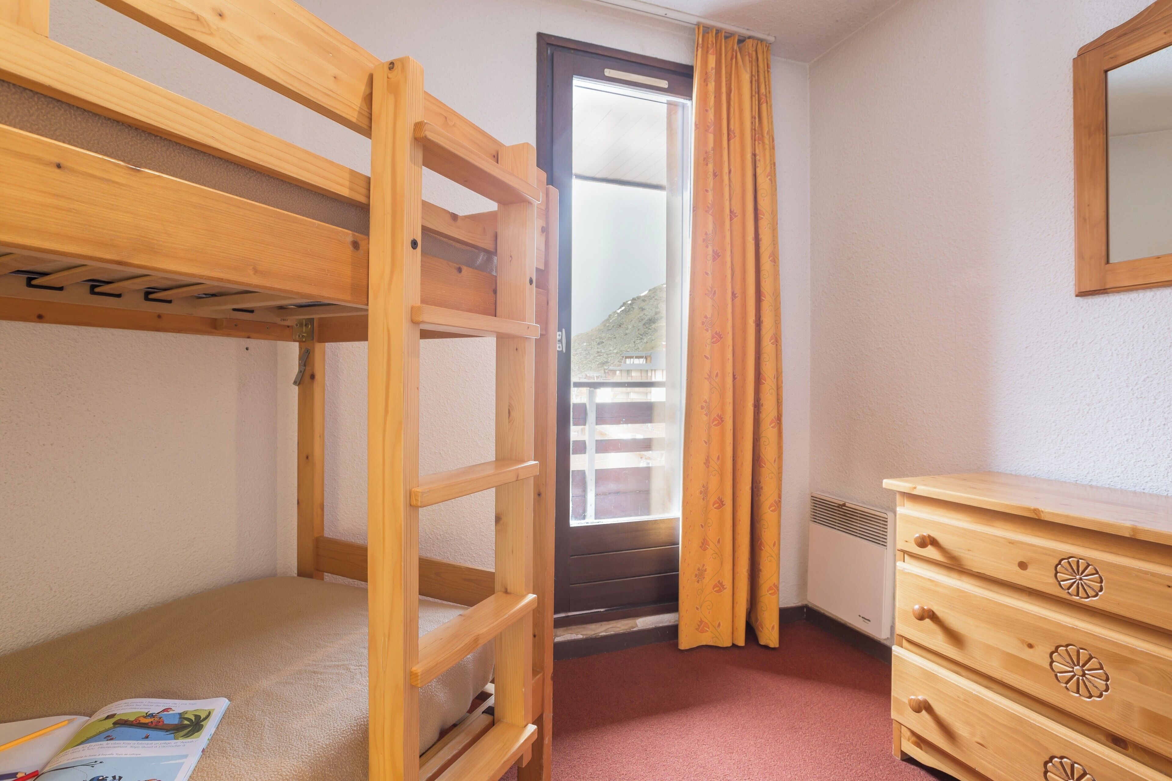 Una habitación con una cama litera, un aparador de madera, una puerta con vistas y una ventana con cortinas.