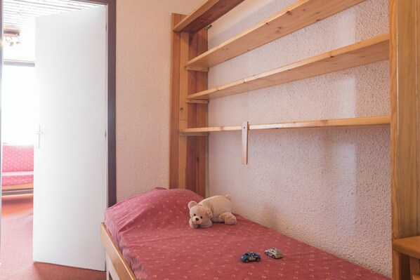A small, single-bed room with a wooden headboard, a red bedspread, and a teddy bear on the bed. - Résidence Le Schuss (Les Belleville)