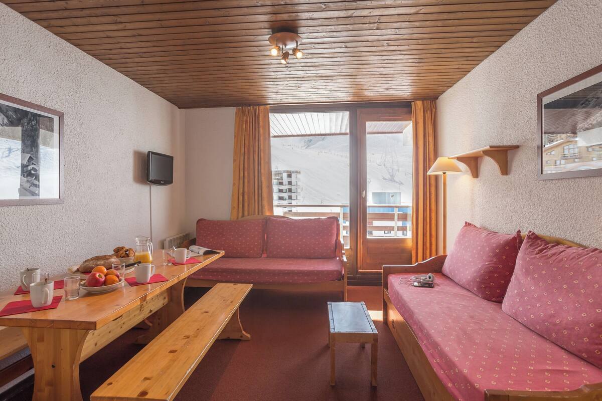 Ein gemütliches Zimmer mit Holzbalkendecke, einer Couch, einer Bank, einem Tisch mit Frühstücksangeboten und einem Fenster mit Blick auf Gebäude.
