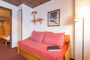 A wooden bed with a red patterned bedspread, a bedside lamp, a framed picture on the wall, and a wooden shelf with flowers and candles. - Résidence Le Schuss (Les Belleville)