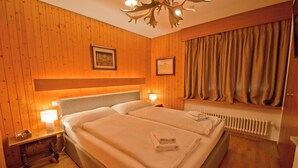 A double bed with a headboard, bedside lamps, a window with curtains, and a radiator. - La Villa Emeline - Wohnung Emilie (Zermatt)