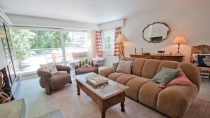 A living room with a sofa, armchair, and coffee table, a dining area with chairs and a table, and a balcony with a view of trees and buildings. - La Villa Emeline - Wohnung Emilie (Zermatt)
