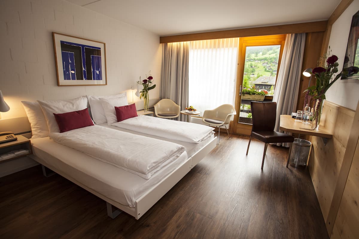A hotel room with two beds, a desk, a chair, a window with a view, and a painting on the wall.