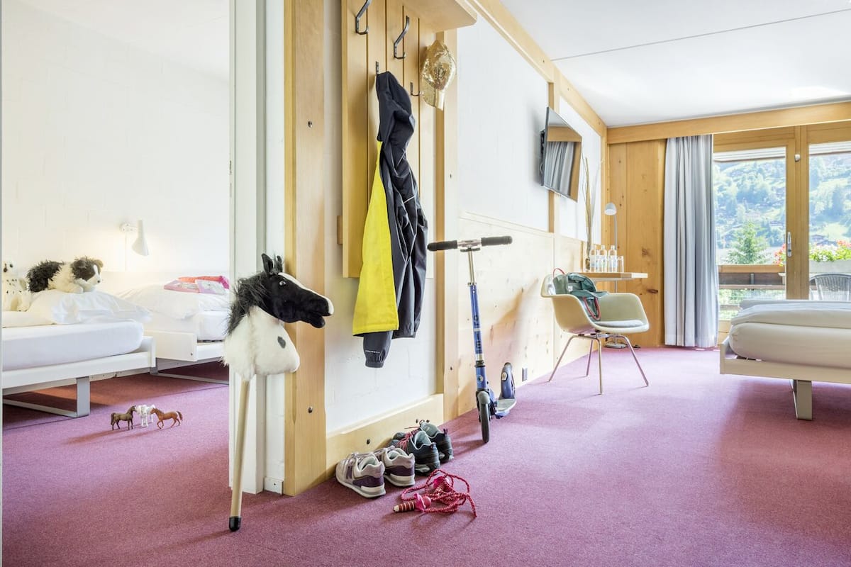 A room with a bed, a desk, a chair, a wall-mounted TV, a window with a view, and a wall with a coat rack and shoes.