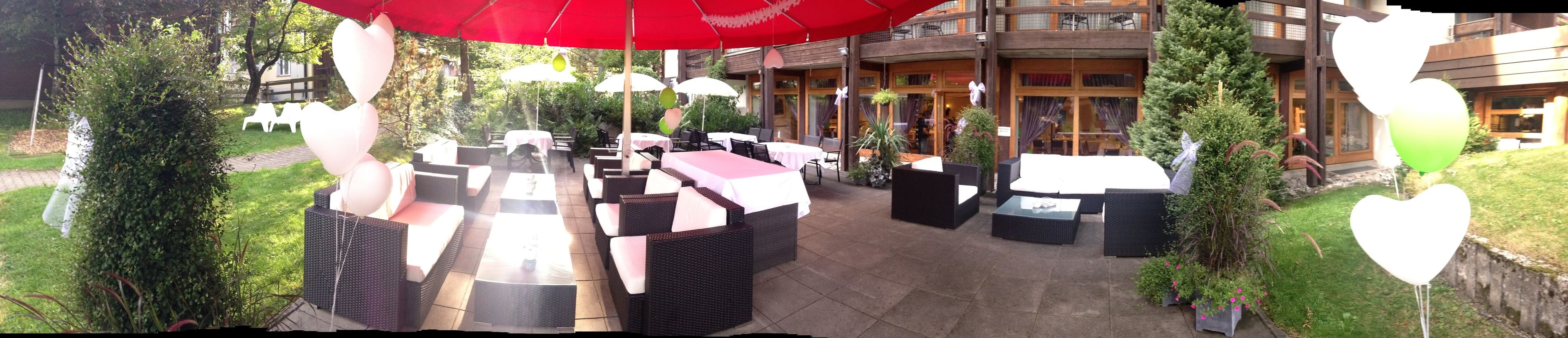 Een terras buiten met rieten meubels, witte kussens en rode parasols.