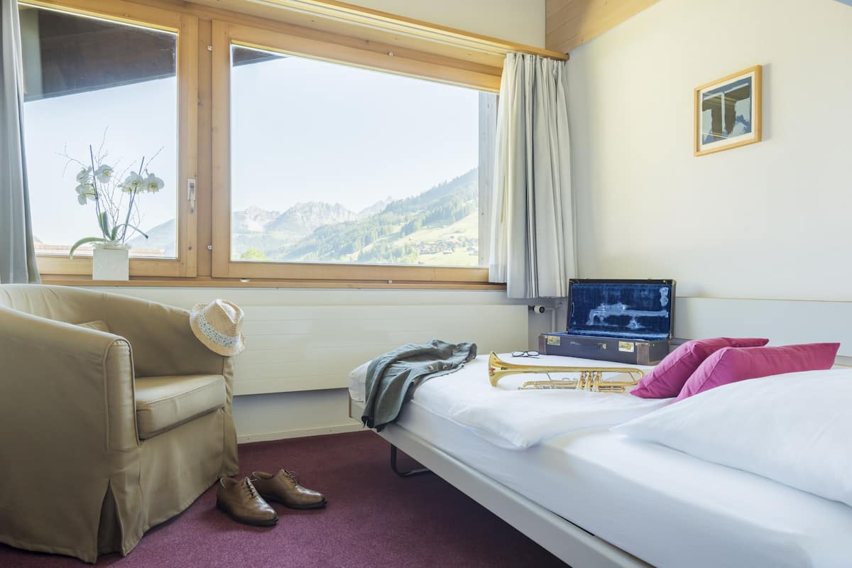 A hotel room with a bed, a chair, a suitcase, and a window with a mountain view.