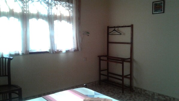 A room with a bed, a chair, a wooden shelf, a window with sheer curtains, and a wall-mounted picture. - Villa Rodrigo (Negombo)