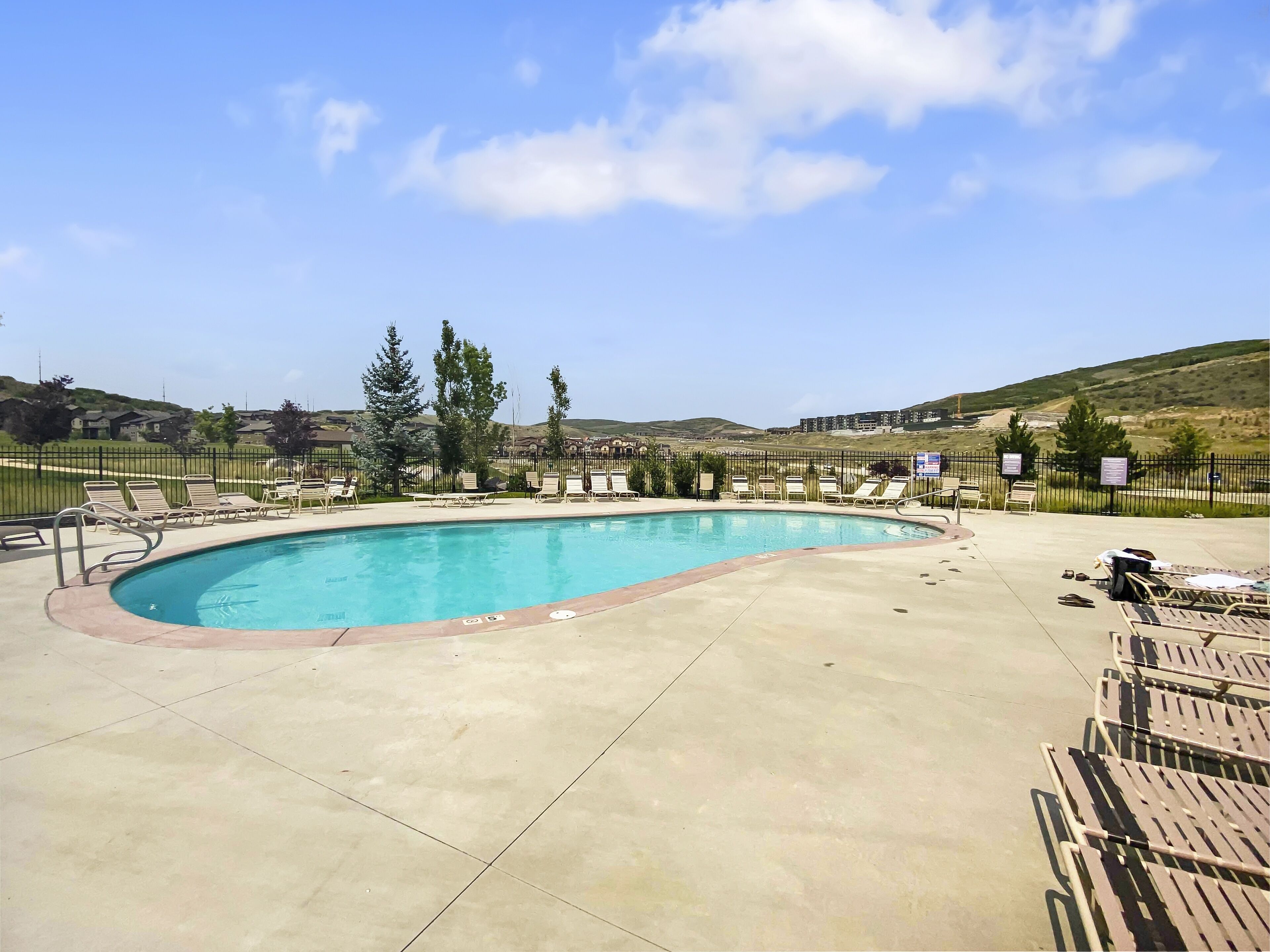 A swimming pool with lounge chairs and a fenced area, surrounded by hills and a clear sky.