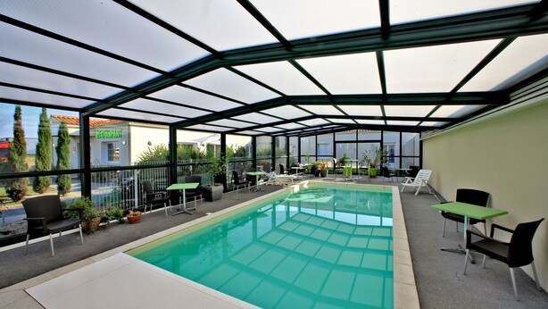 An indoor swimming pool with a glass ceiling, surrounded by lounge chairs and tables, and a restaurant in the background. - Logis L'auberge de Moricq (Angles)
