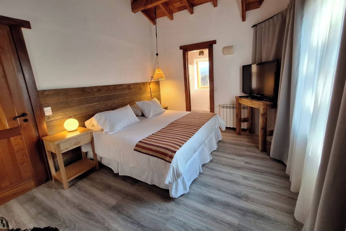 Une chambre à coucher avec une tête de lit en bois, un lit avec une literie blanche, une lampe de chevet, un téléviseur et une fenêtre avec des rideaux.