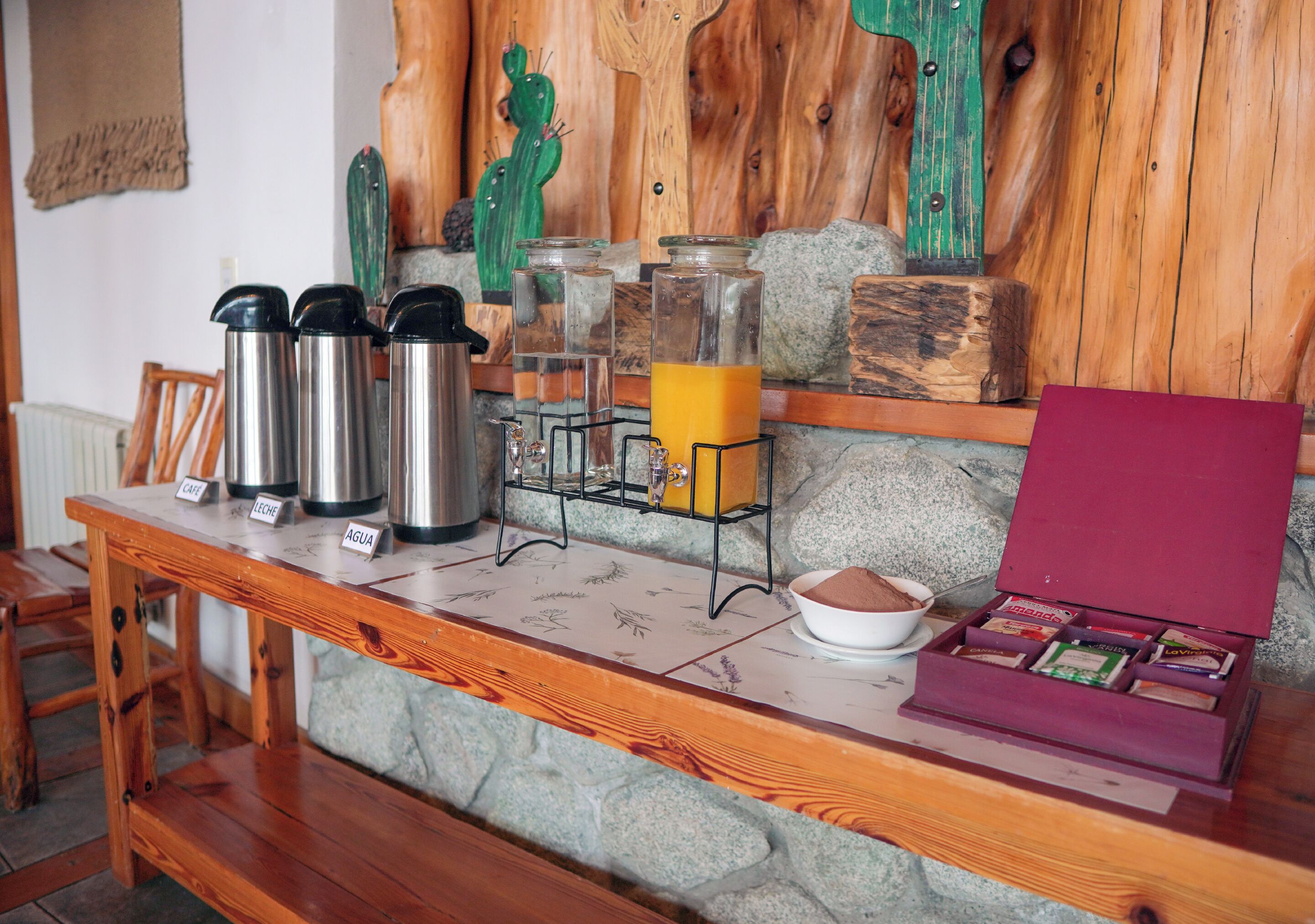 A breakfast bar with stainless steel thermos cups labeled 'Café' and 'Agua', a juice dispenser, and a box of assorted pastries.