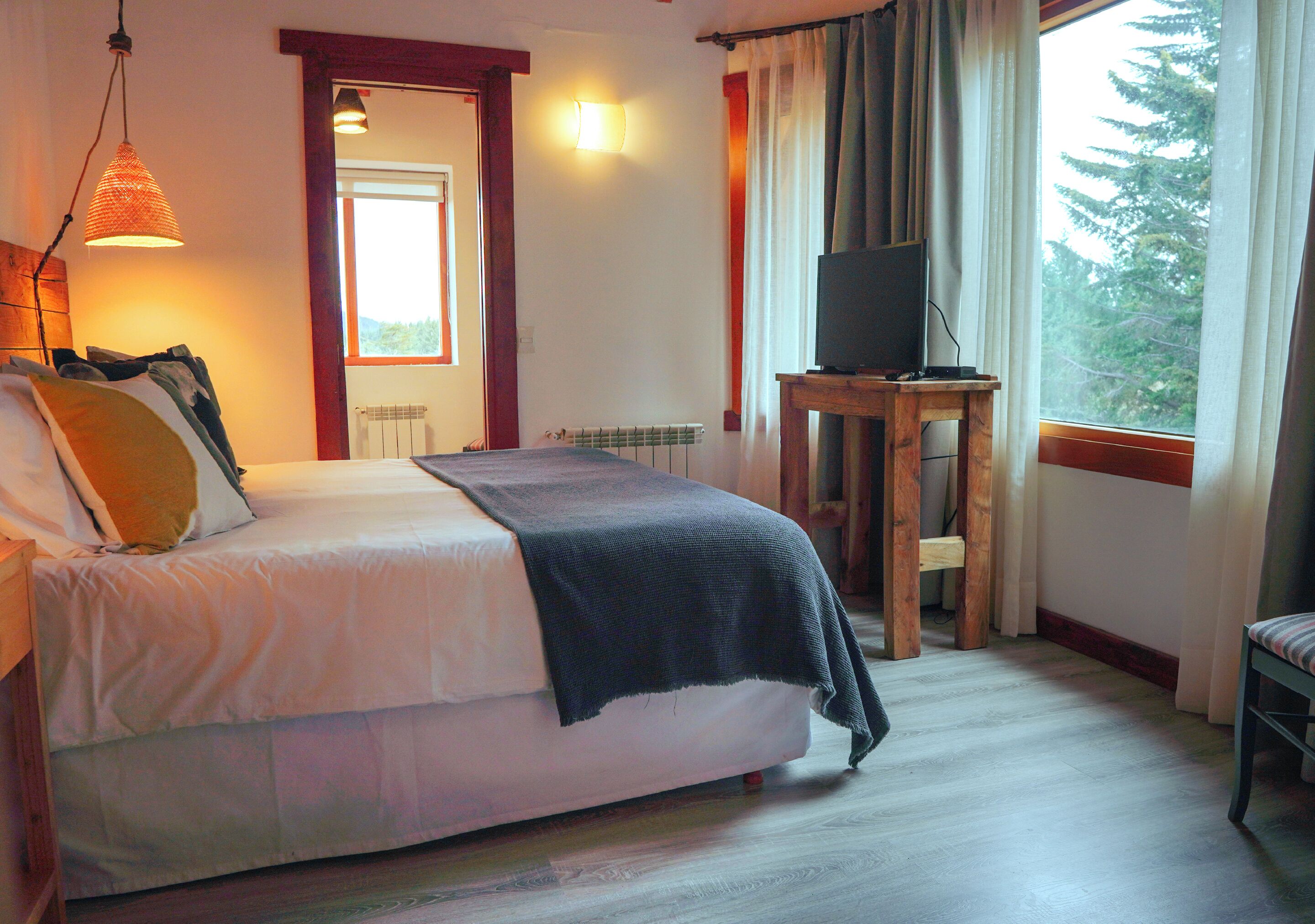 A bedroom with a bed, a TV on a wooden table, a window with curtains, and a view of trees outside.