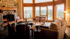 A cozy room with a stone fireplace, wooden furniture, and large windows offering a view of the outdoors. - El Bosque by DOT Tradition (Villa La Angostura)