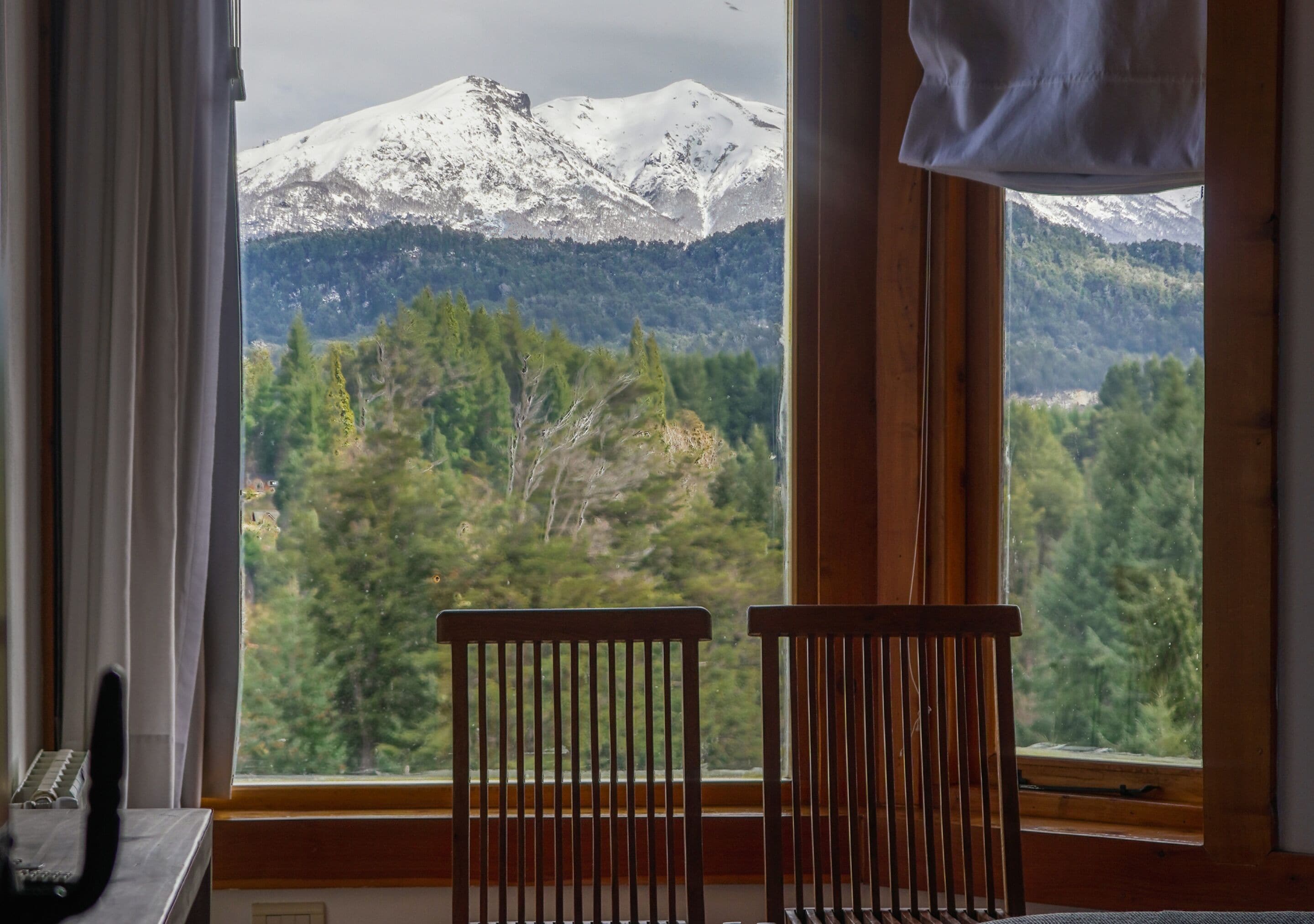 A room with a view of a snow-capped mountain and forest. There are two wooden chairs in front of a window.