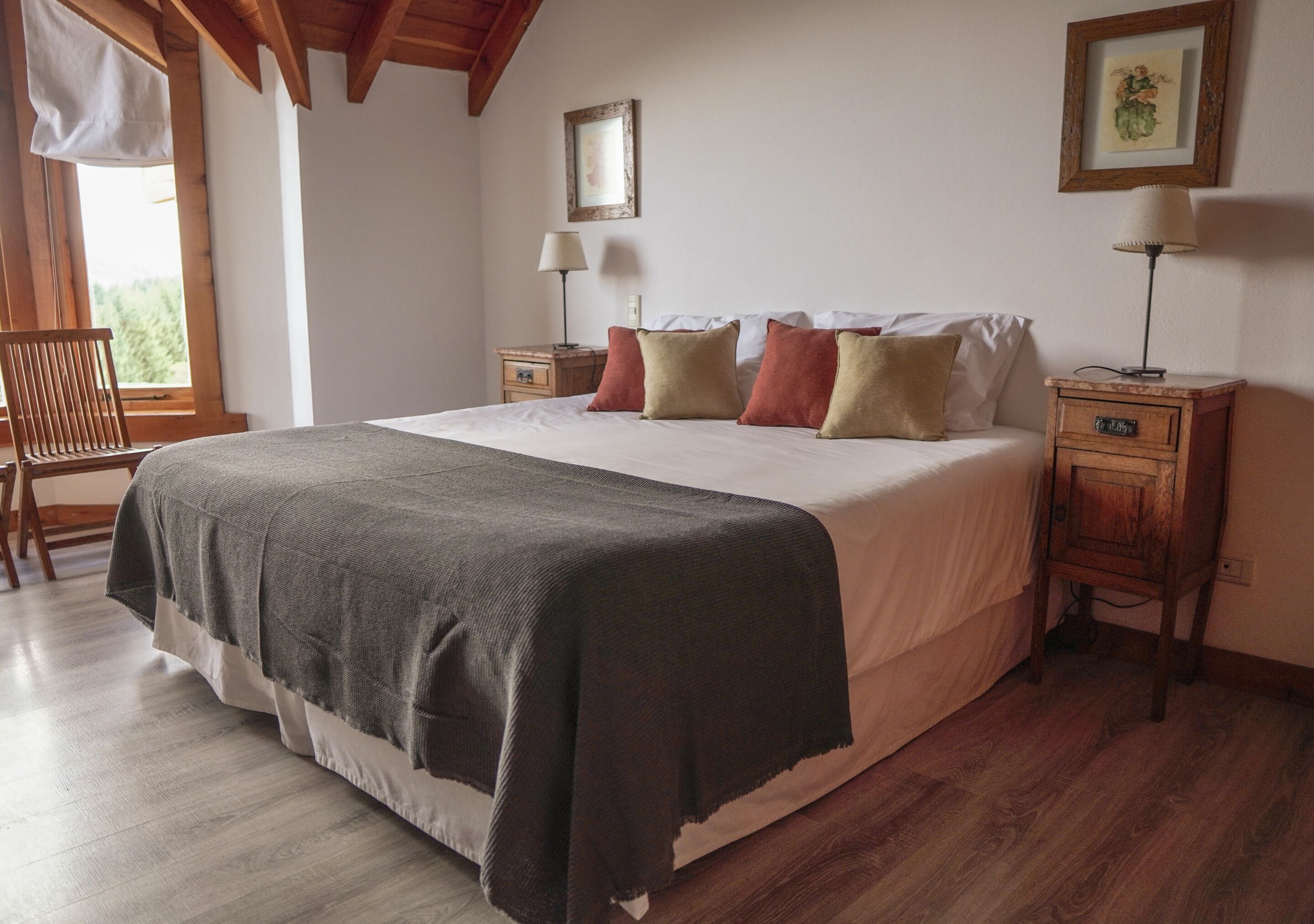 Un dormitorio con una cama grande, cabecera de madera, dos mesitas de noche con lámparas, una silla y una ventana con vistas a la vegetación.