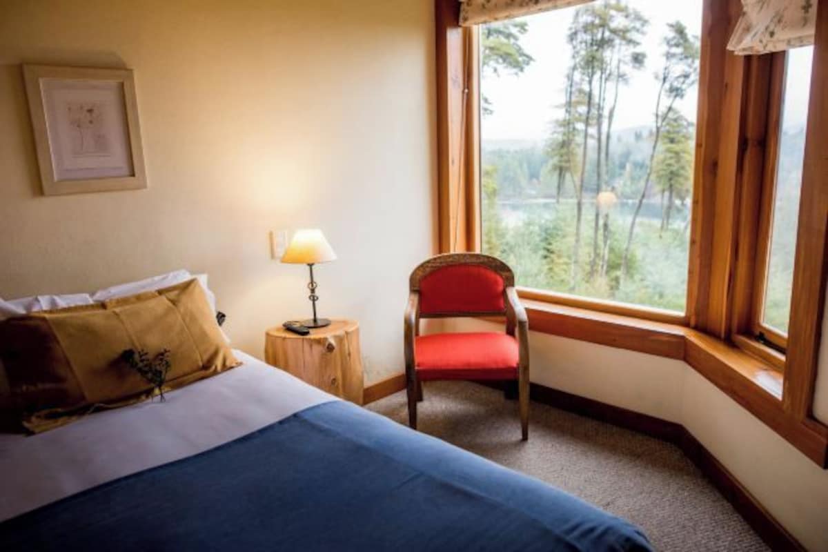 A bedroom with a bed, a chair, a nightstand, a lamp, and a window with a view of trees.