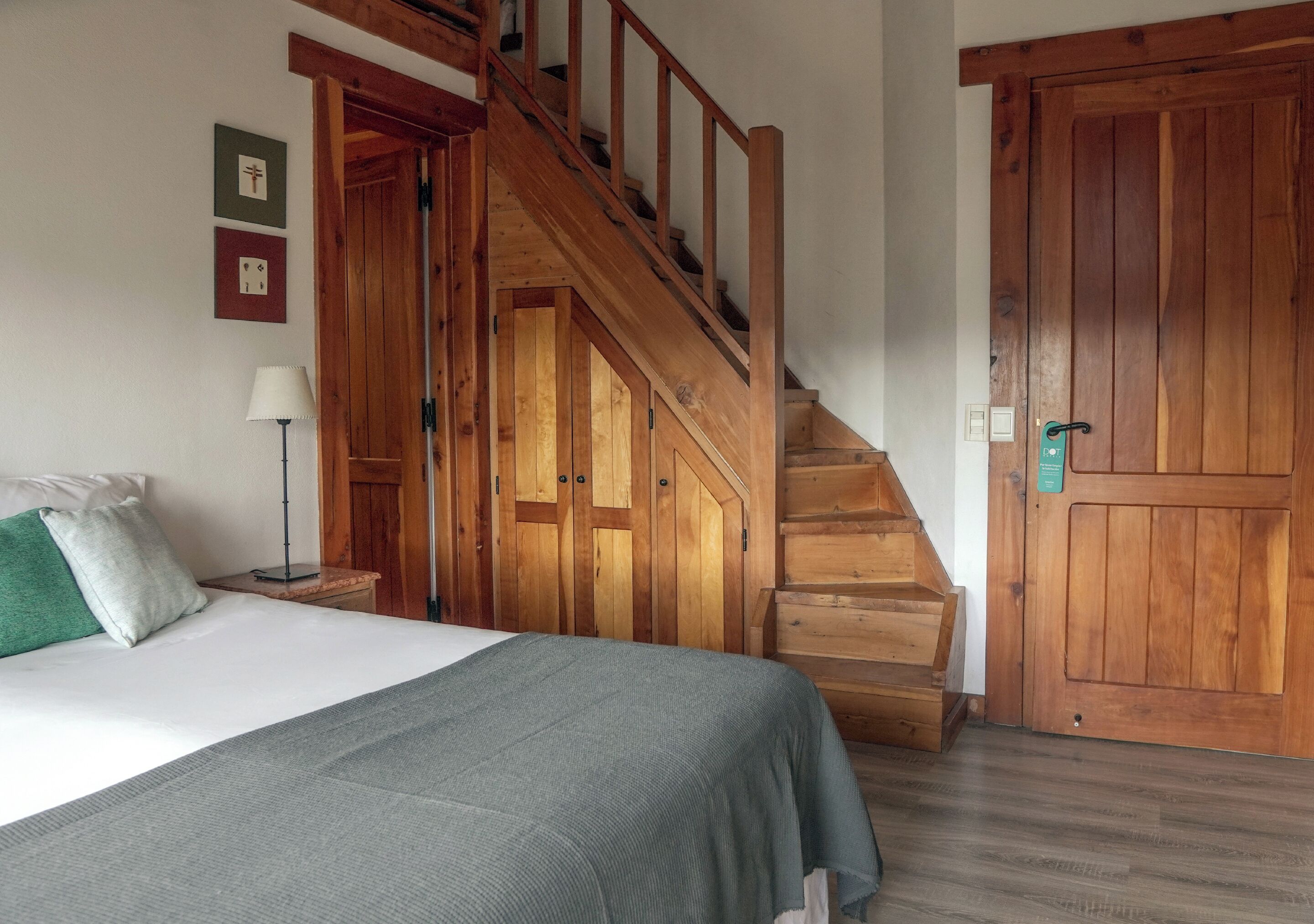 Un dormitorio con una escalera de madera, una cama con un edredón gris, una mesita de noche con una lámpara y dos cuadros en la pared.