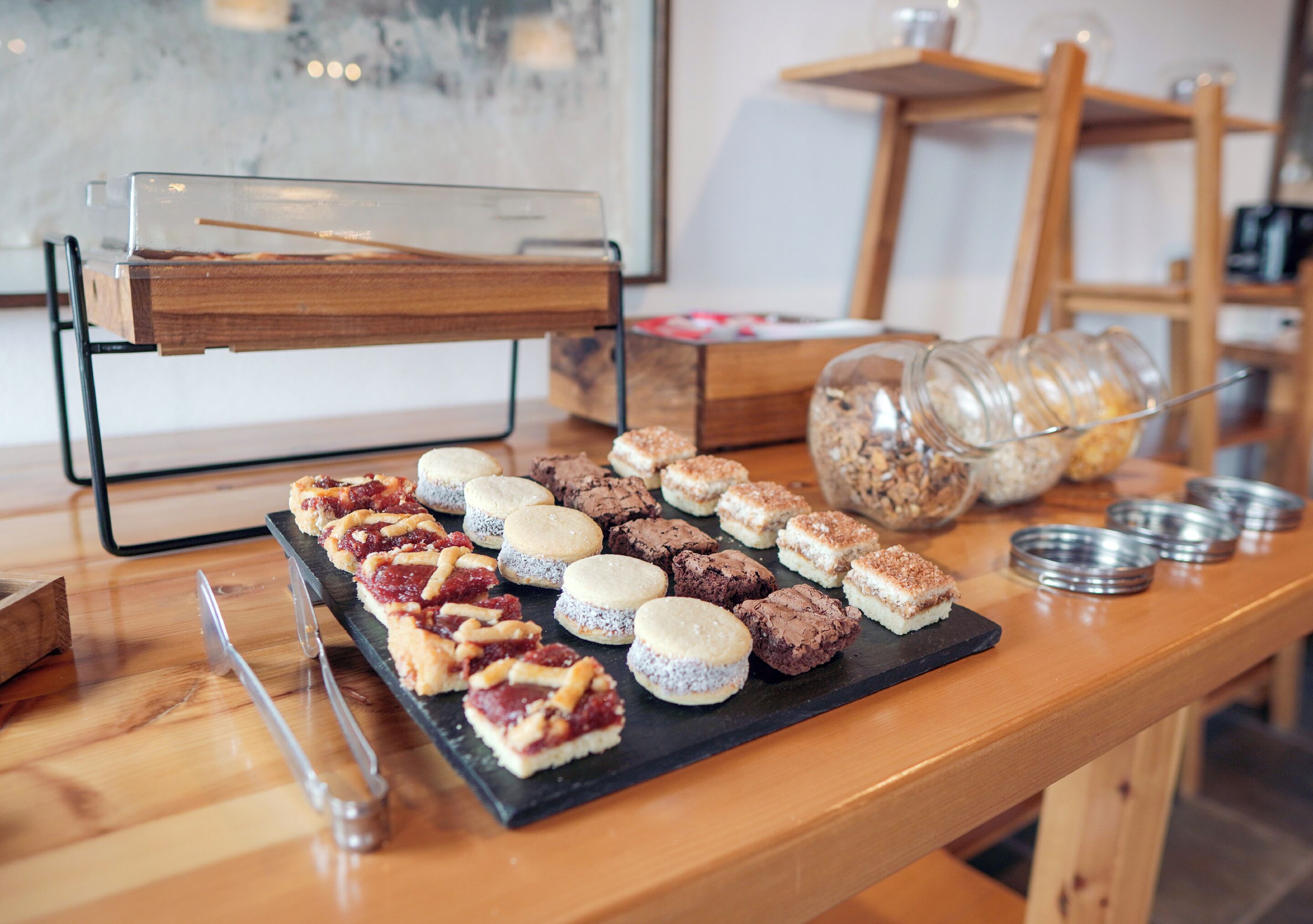 Ett urval av bakverk och desserter på ett träbord, med ett glasskåp och metallbehållare i bakgrunden.