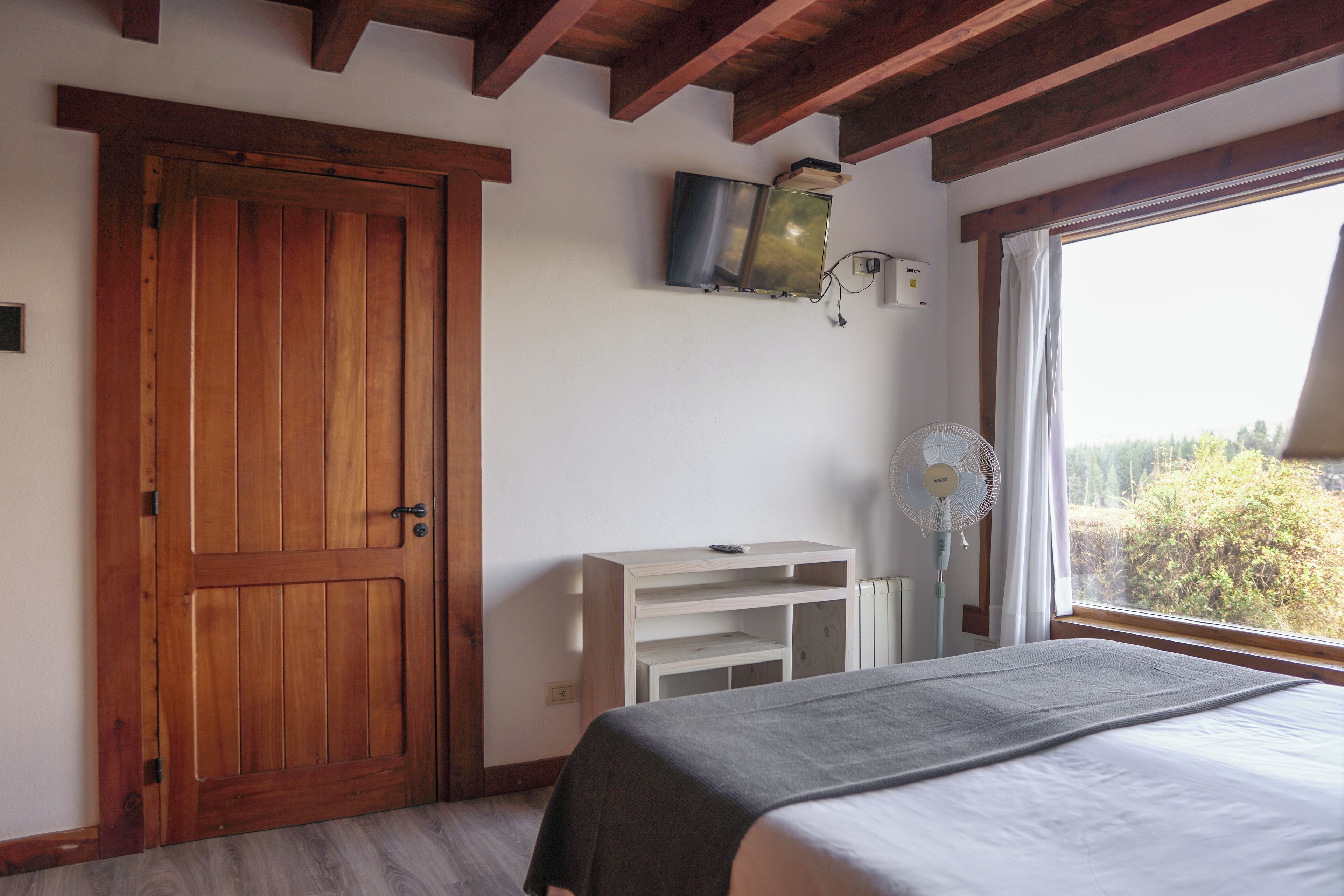 Un dormitorio con puerta de madera, cama con manta gris, mesita de noche blanca, televisor montado en la pared y ventana con vistas a árboles.