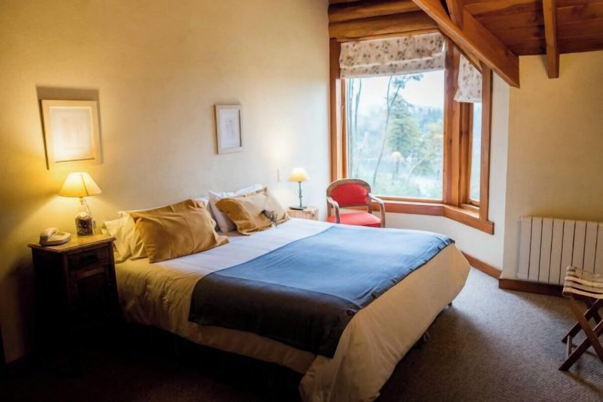 A bedroom with a bed, bedside lamps, a chair, a window with a view of trees, and a radiator.