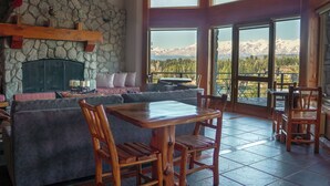 A cozy interior with a stone fireplace, a wooden dining table, and a large window offering a mountain view. - El Bosque by DOT Tradition (Villa La Angostura)