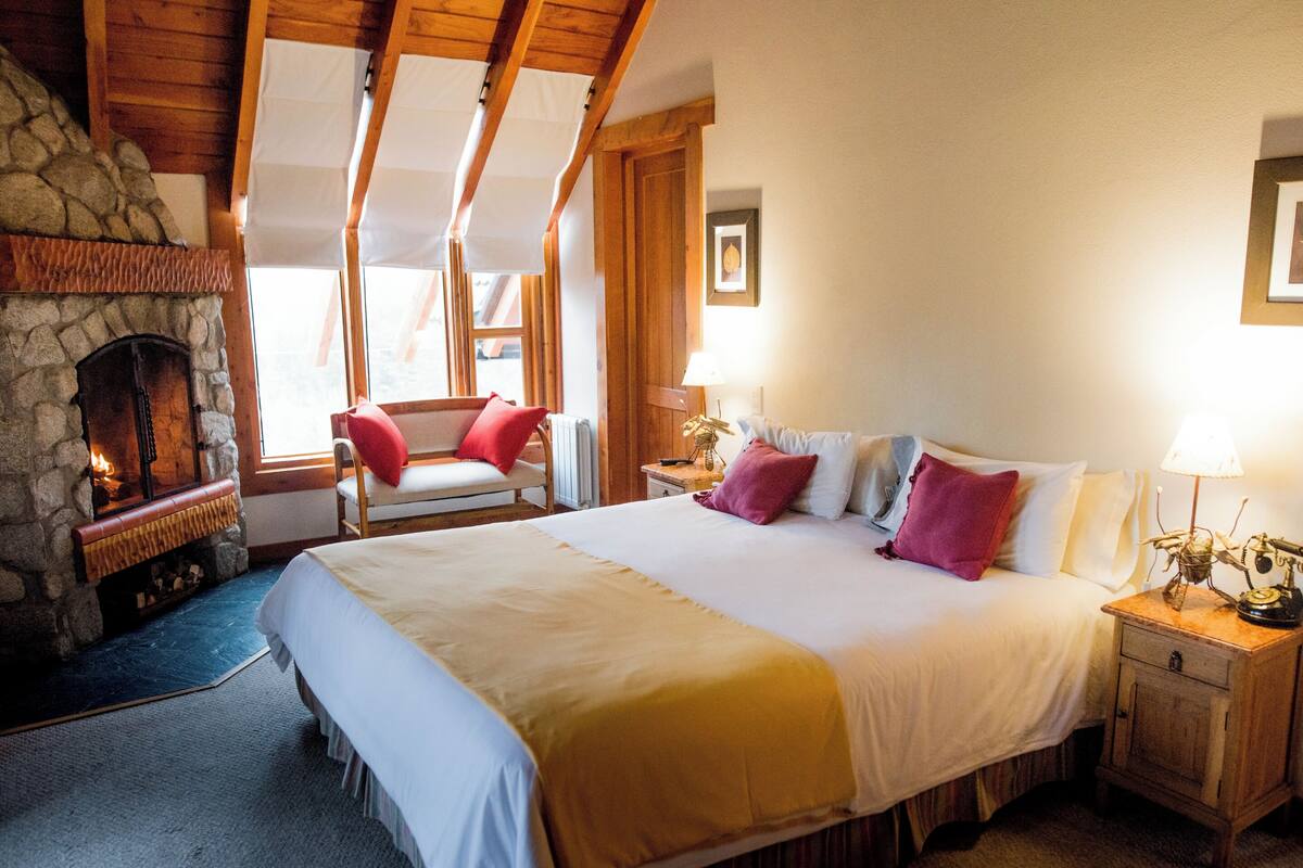 Une chambre confortable avec une cheminée en pierre, un grand lit agrémenté de nombreux coussins, une table de chevet en bois et un fauteuil près de la fenêtre.