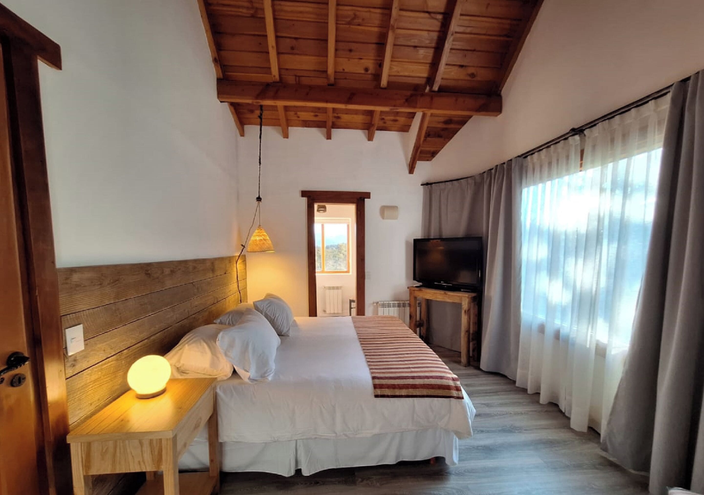 Ein Schlafzimmer mit Holzboden, einem Bett mit weißer Bettwäsche, einer Nachttischlampe, einem Fernseher auf einem Holztisch und einem Fenster mit Vorhängen.