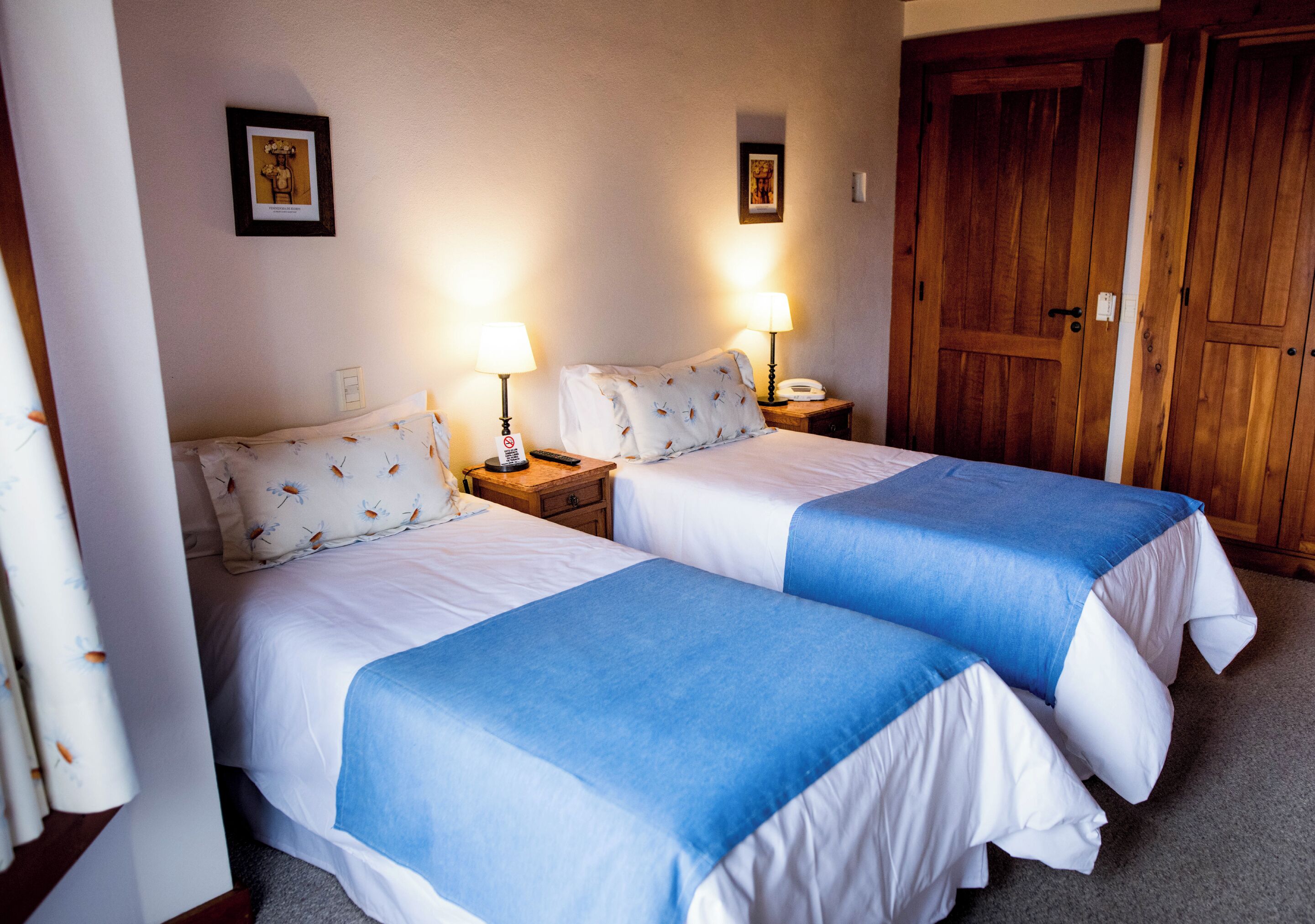 Habitación de hotel con dos camas, cada una con colcha azul y blanca, lámparas de noche y un armario de madera.